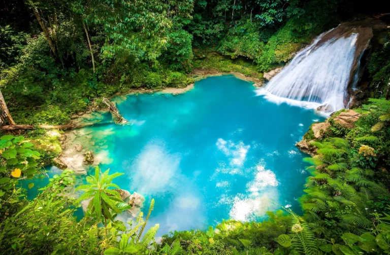 Cascata a desaguar num lago azul-turquesa rodeado de vegetação tropical densa na Jamaica, refletindo a natureza exuberante e os cenários paradisíacos da ilha caribenha.