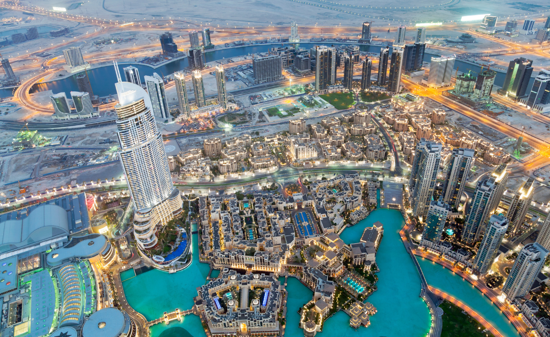 Vista aérea de Downtown Dubai, mostrando arranha-céus iluminados, o lago turquesa junto ao Dubai Mall e zonas residenciais modernas rodeadas por vias rápidas.