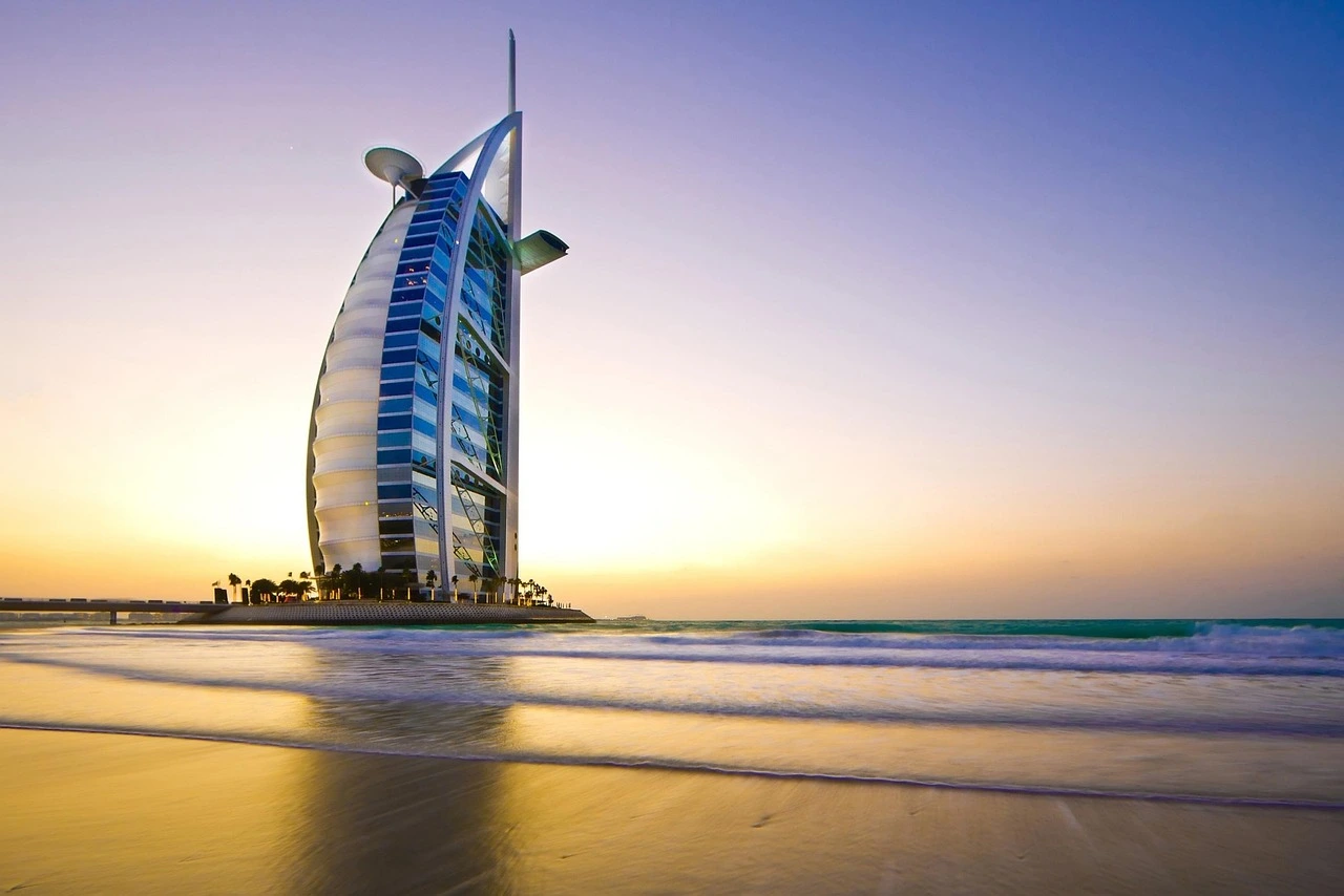 Burj Al Arab iluminado pelo pôr do sol, visto a partir da praia com reflexo dourado na água e céu em tons pastel