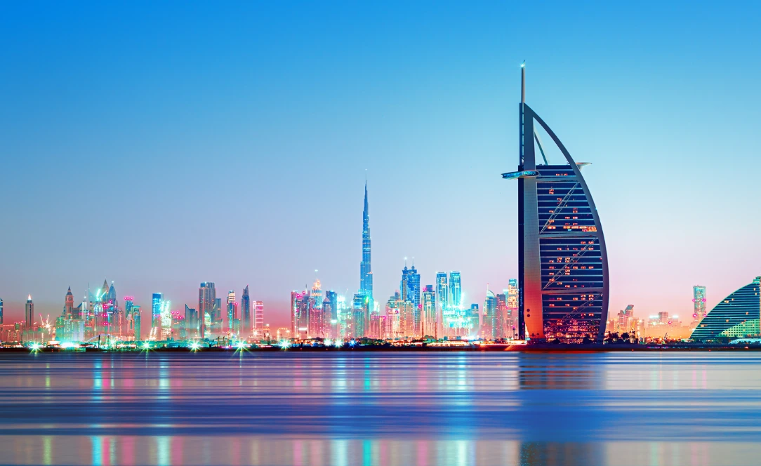Vista do Burj Al Arab iluminado ao entardecer, com o skyline moderno de Dubai e o Burj Khalifa ao fundo refletidos nas águas calmas.