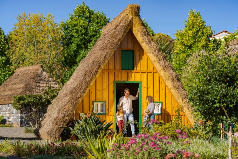 Família a visitar uma casa típica de Santana, na Madeira, com estrutura em madeira e cobertura de colmo, rodeada por jardins floridos num dia solarengo.