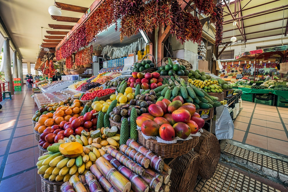 Banca colorida no Mercado dos Lavradores, no Funchal, repleta de frutas tropicais frescas como mangas, bananas, anonas e maracujás, com ambiente tradicional madeirense.