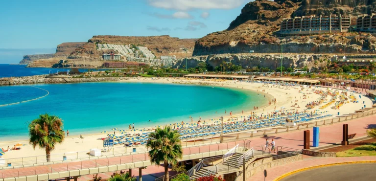 Vista panorâmica da Praia de Amadores, em Gran Canaria, com águas turquesa, areia dourada, espreguiçadeiras e falésias ao fundo num dia ensolarado.