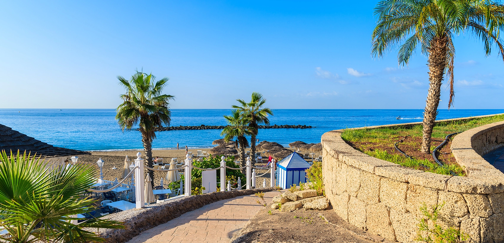 Caminho ladeado por palmeiras que conduz a uma praia tranquila em Gran Canaria, com mar calmo, areia dourada e um pequeno abrigo azul e branco.