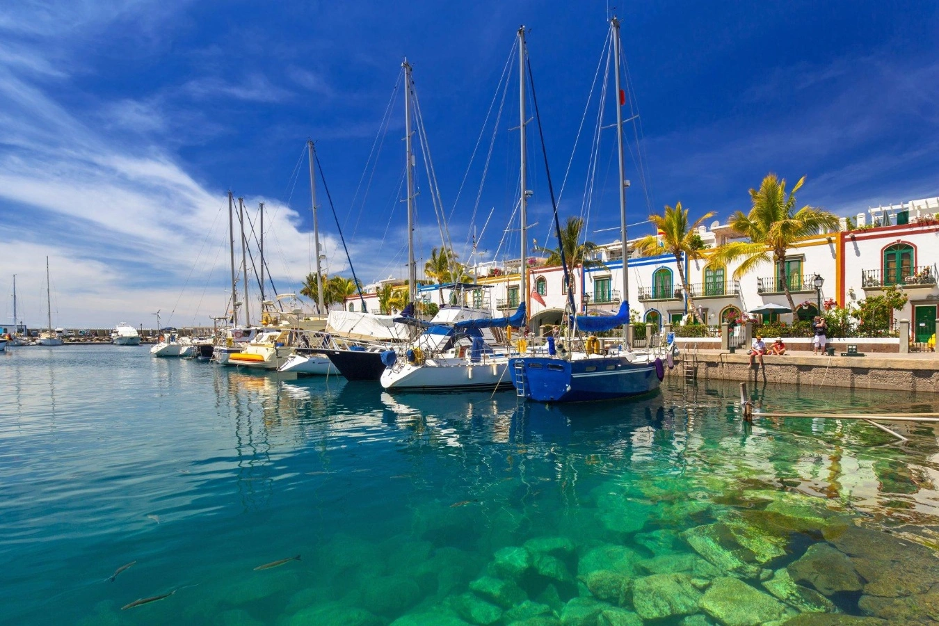 Porto de Mogán, em Gran Canaria, com barcos atracados sobre águas turquesa cristalinas e casas coloridas com palmeiras ao fundo.