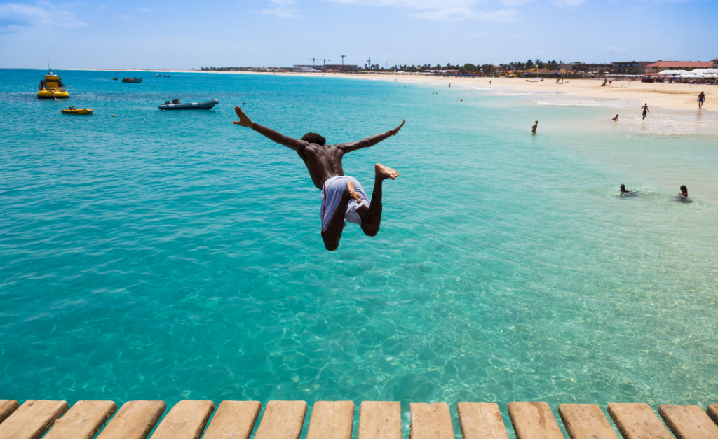 Rapaz a saltar de um pontão para o mar turquesa da Ilha do Sal, com praia e barcos ao fundo.