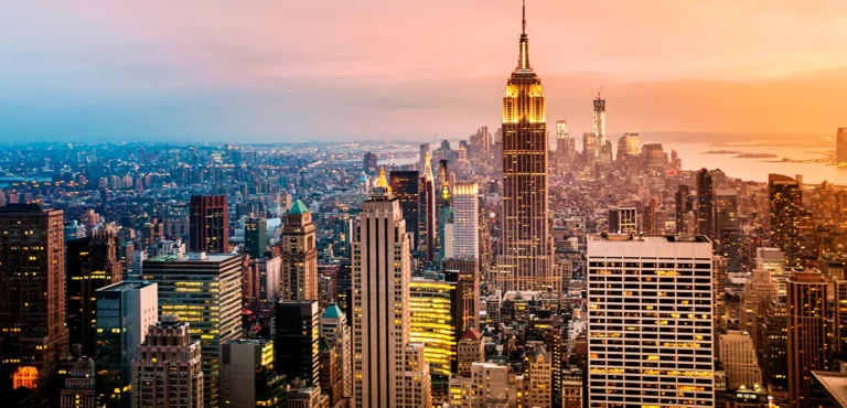 Vista panorâmica de Manhattan ao pôr do sol, com o Empire State Building iluminado no centro e arranha-céus a perder de vista.