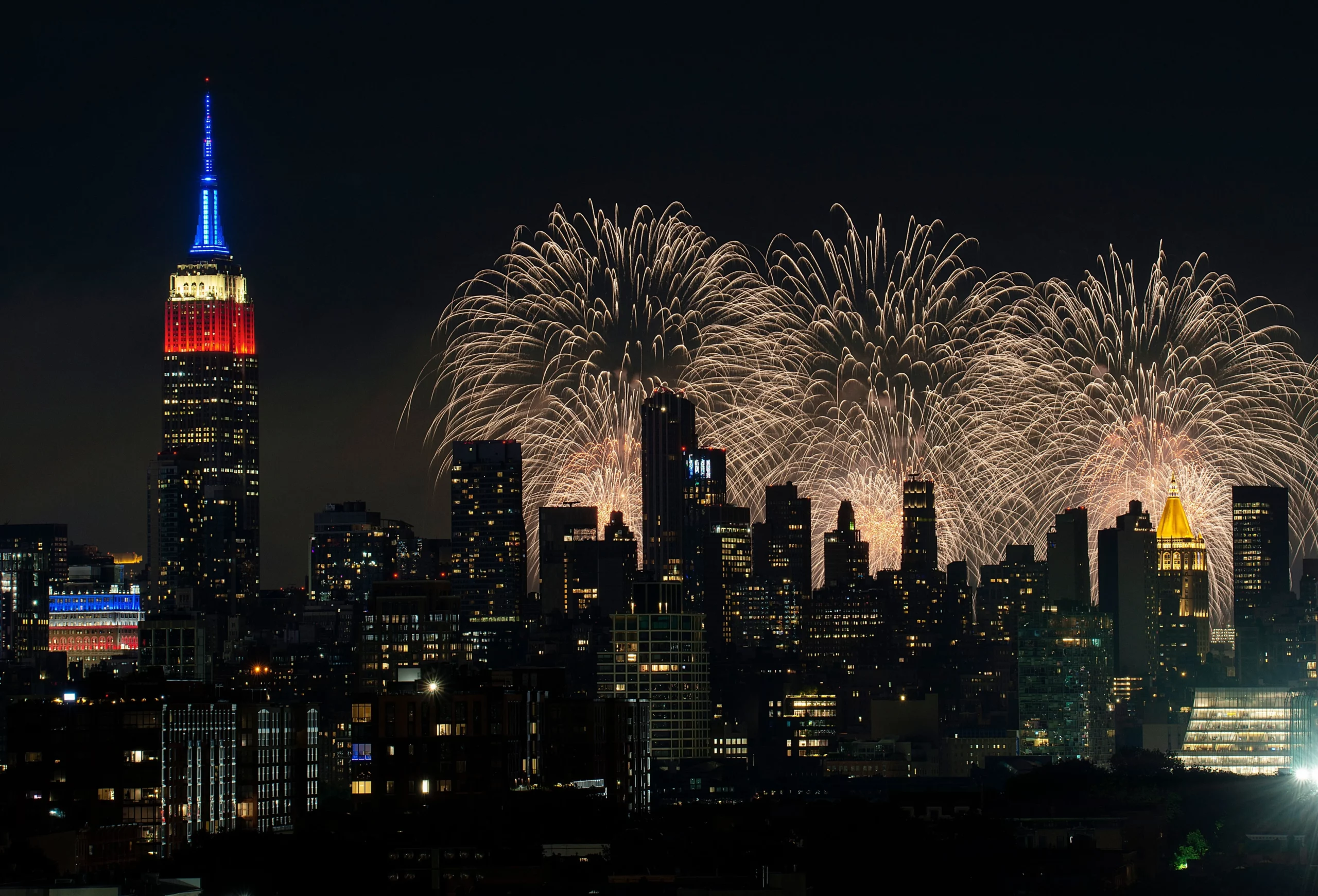 Show de fogo de artifício iluminando o céu noturno de Nova Iorque, com o Empire State Building aceso em cores vibrantes ao lado dos arranha-céus.