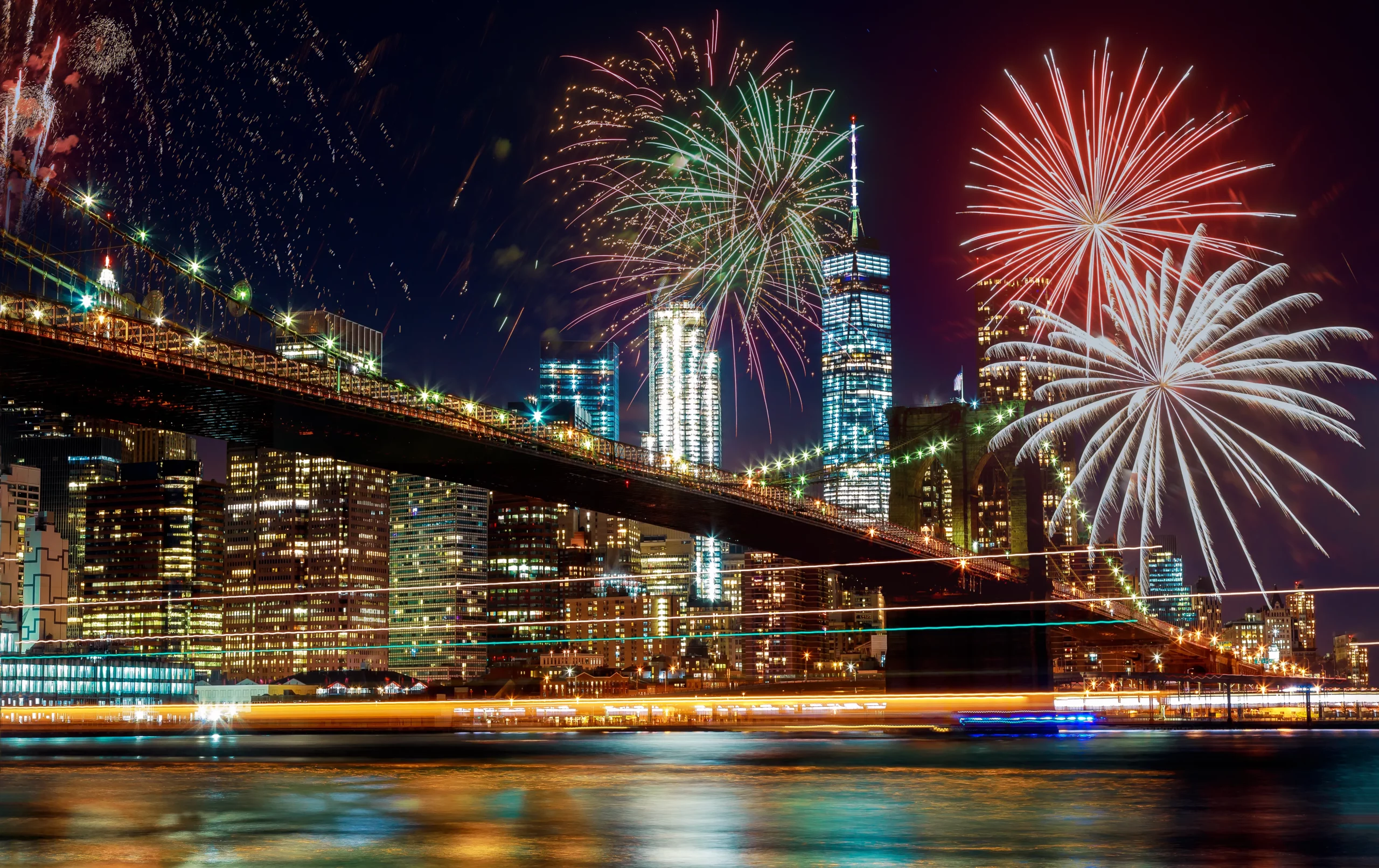 Fogo de artifício colorido a iluminar o céu noturno de Nova Iorque, com a Ponte de Brooklyn e os arranha-céus refletidos na água.