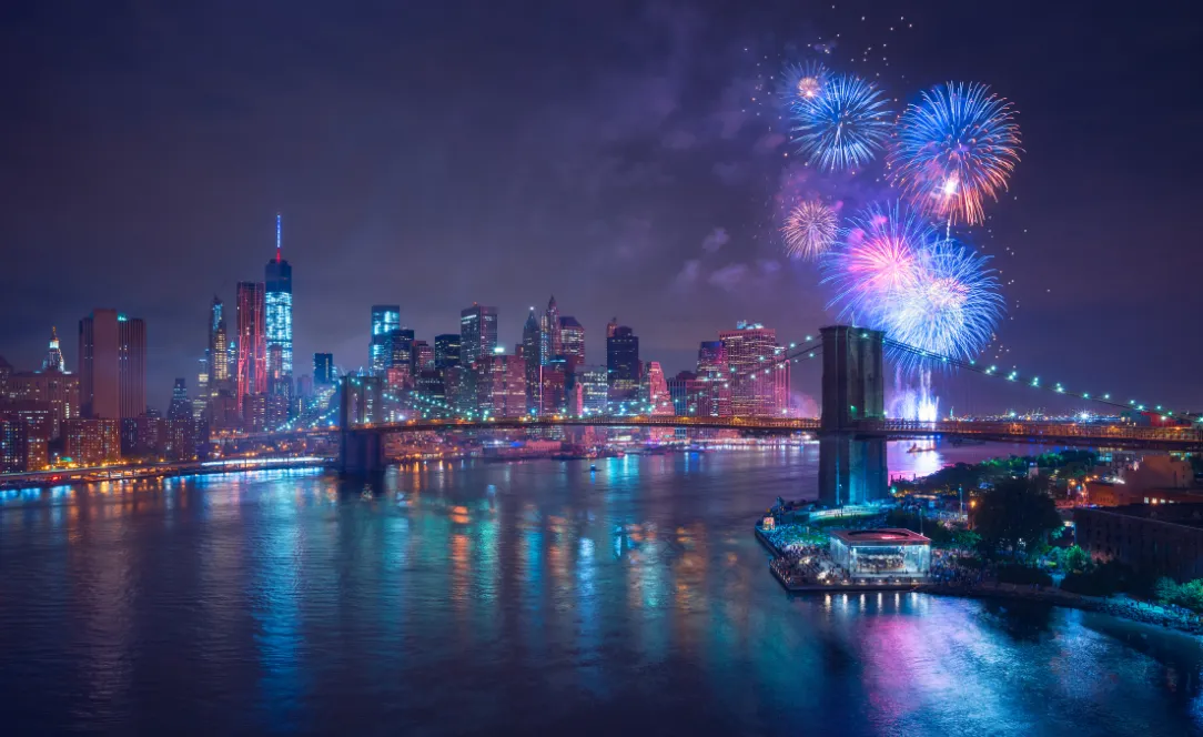 Vista noturna de Manhattan refletida no rio, com fogo de artifício colorido a iluminar o céu e a Ponte de Brooklyn ao lado.