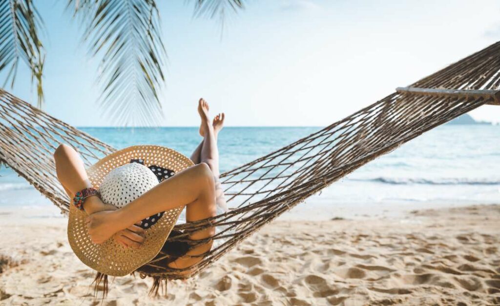 Mulher de chapéu de palha deitada numa rede à beira-mar na Riviera Maya, com areia dourada e mar calmo ao fundo.