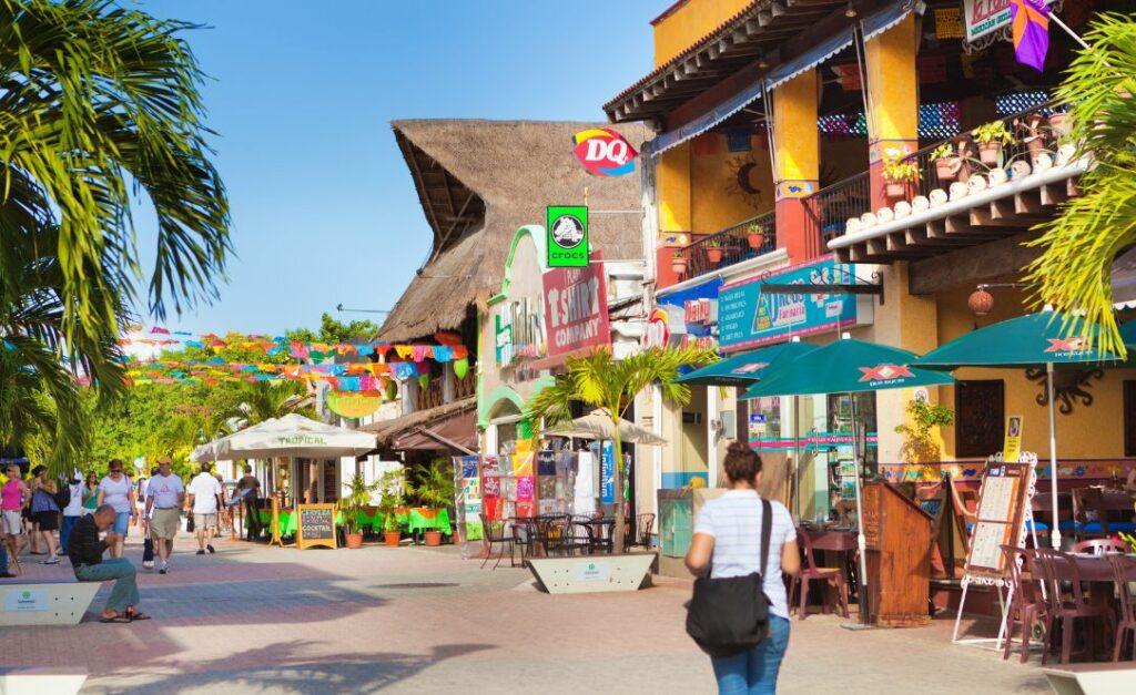 Rua movimentada da Riviera Maya com lojas coloridas, restaurantes, decoração com bandeirolas e turistas a passear.