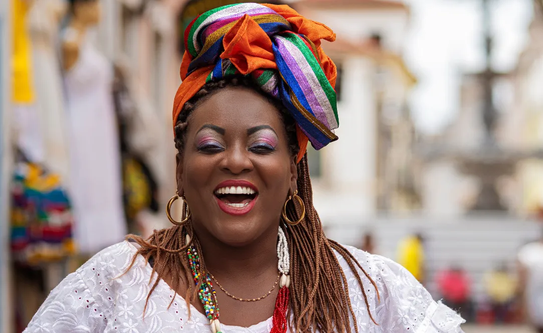 Mulher baiana vestida com traje tradicional colorido, usando turbante vibrante e colares, sorrindo nas ruas históricas do Pelourinho em Salvador, Bahia.