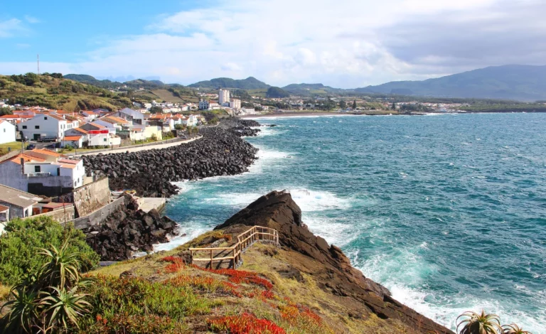 Vista costeira da Ribeira Grande, em São Miguel, Açores, mostrando as casas junto ao mar, as rochas vulcânicas negras, o miradouro sobre a falésia e o oceano Atlântico ao fundo.