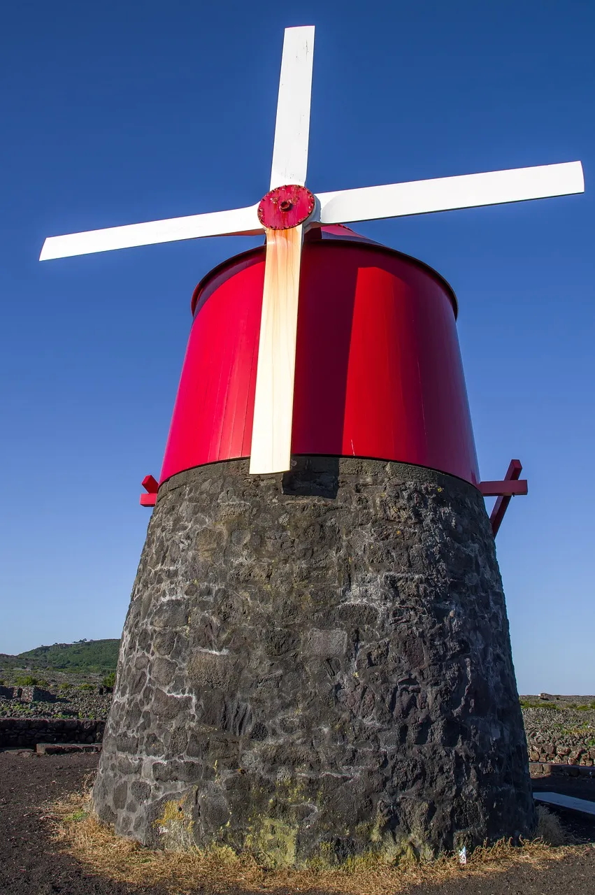 Moinho açoriano com base de pedra vulcânica e corpo vermelho, com pás brancas, fotografado sob um céu azul limpo.