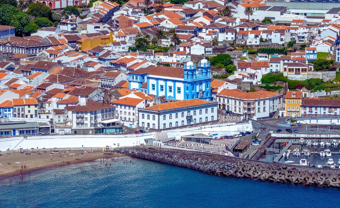 Vista aérea do centro histórico de Angra do Heroísmo, mostrando casas de telhados laranja junto ao mar e a Igreja da Conceição em azul e branco em posição central.