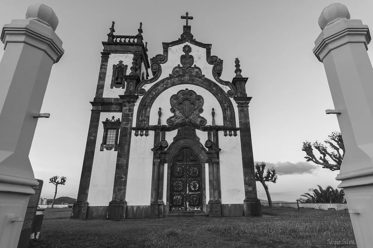 Vista frontal de uma ermida ornamentada na Ilha Terceira, com arco central detalhado e cruz no topo, captada em preto e branco ao entardecer.