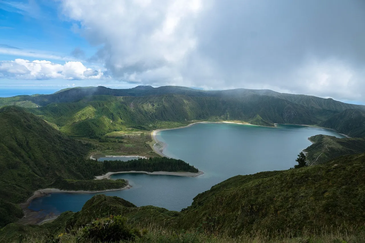 Paisagem panorâmica da Lagoa do Fogo, nos Açores, com colinas verdes, céu parcialmente nublado e águas azul-turquesa ao centro.