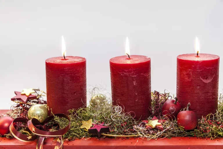 Três velas vermelhas acesas sobre uma decoração natalícia com ramos verdes, bagas, laços e pequenos ornamentos dourados.