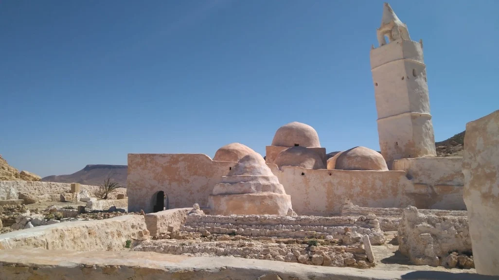 Construções tradicionais de cor areia com cúpulas e torre antiga em Djerba, Tunísia, sob um céu azul claro.