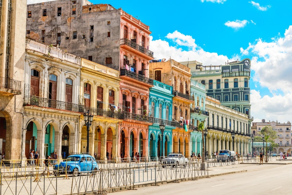 Prédios coloniais coloridos ao longo de uma rua emblemática de Havana, Cuba, com fachadas vibrantes e carros clássicos ao fundo.