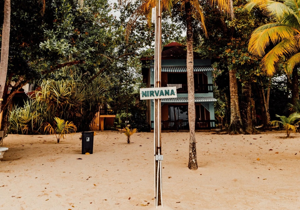 Placa “Nirvana” numa praia tropical com areia dourada, palmeiras e um pequeno alojamento ao fundo