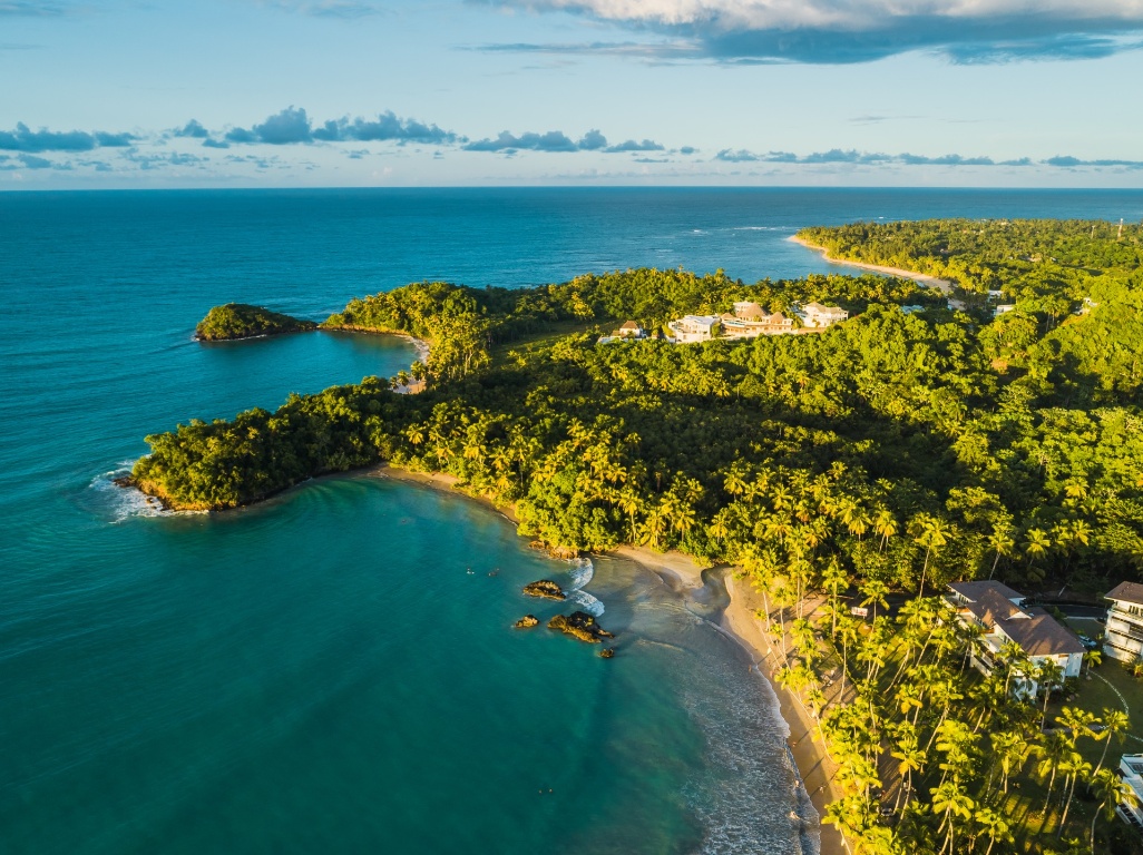 Vista aérea de uma península tropical com praias de água azul-turquesa, vegetação densa e resorts junto à costa ao pôr do sol.