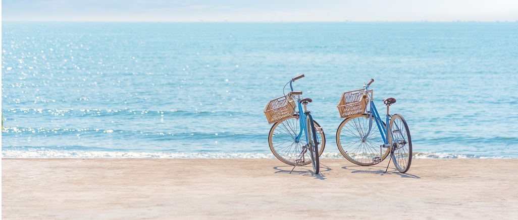 Duas bicicletas azuis com cestos estacionadas junto ao mar calmo numa esplanada à beira-praia.