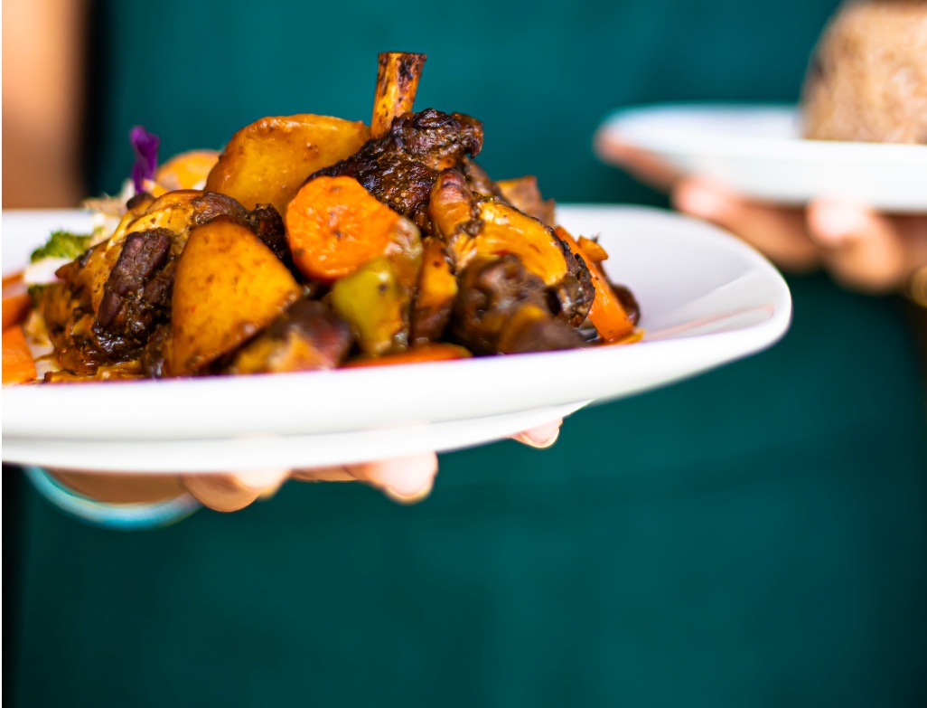 Prato de comida quente com legumes salteados e carne estufada, servido num prato branco segurado por um funcionário de restaurante.