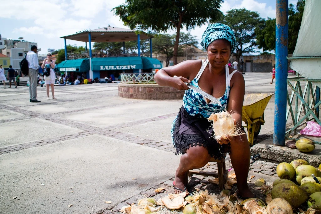 Mulher cabo-verdiana a abrir um coco com faca numa praça da Ilha do Sal, com turistas ao fundo.