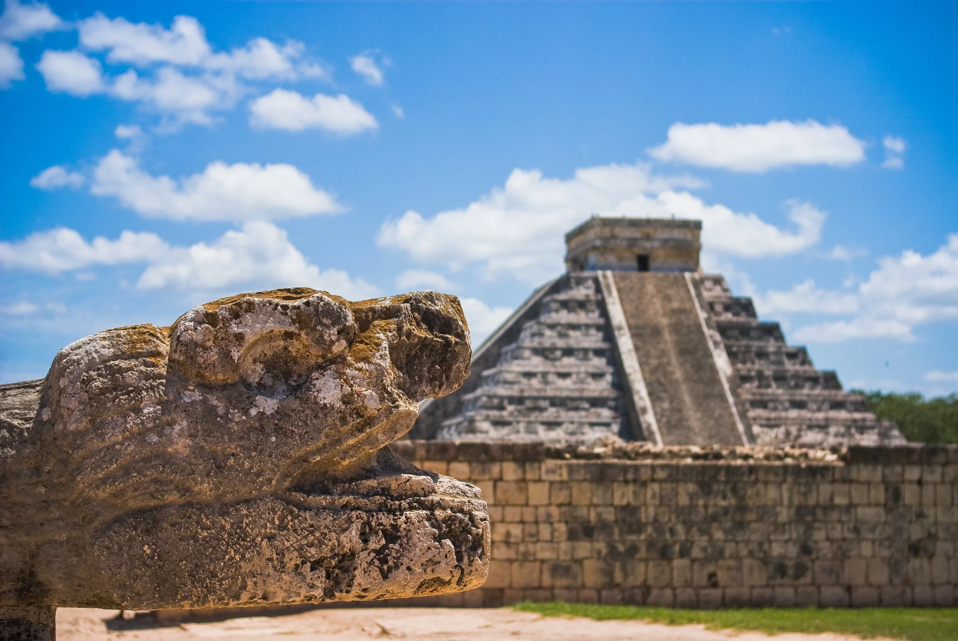 Escultura de serpente em pedra em primeiro plano com a pirâmide de Chichén Itzá ao fundo sob céu azul com nuvens.