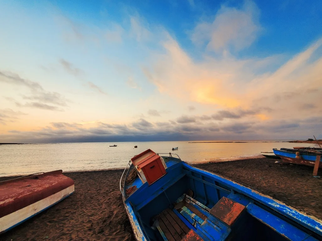 Barcos de pesca coloridos encostados na areia escura da Ilha do Sal, com pôr do sol sobre o mar calmo.