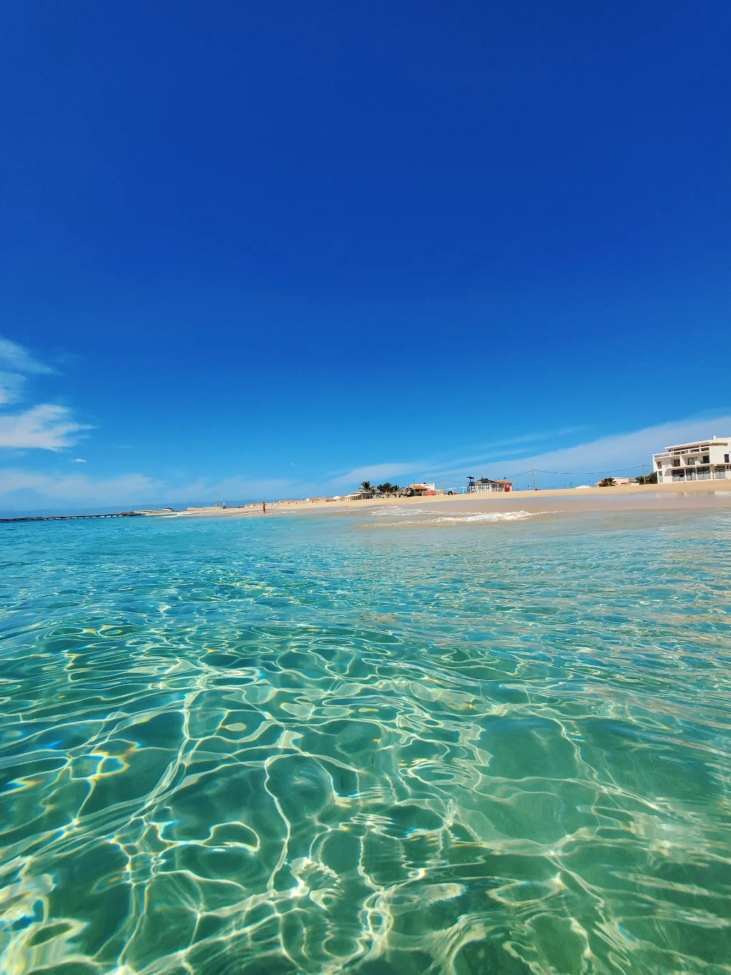 Água extremamente transparente da Ilha do Sal com reflexos de luz, praia de areia clara ao fundo e céu azul intenso.